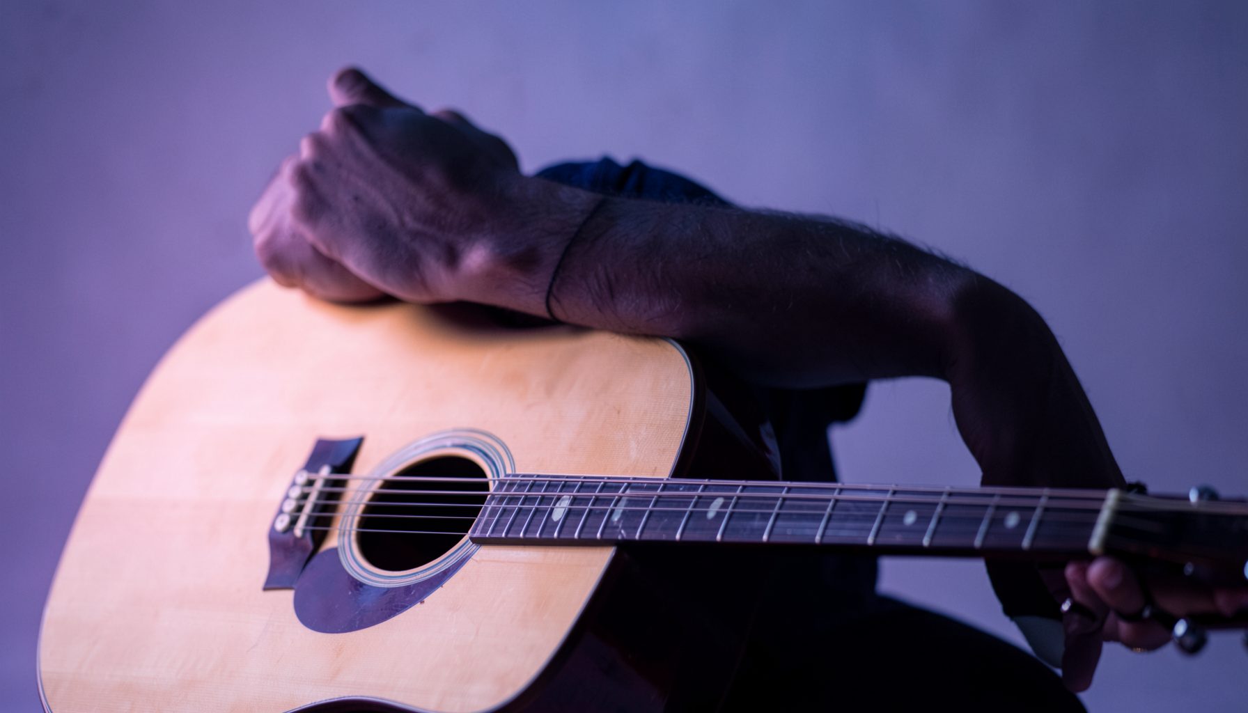 Braço apoiado sobre um violão acústico em madeira clara, iluminação suave em tons roxos e azuis, clima intimista de estúdio