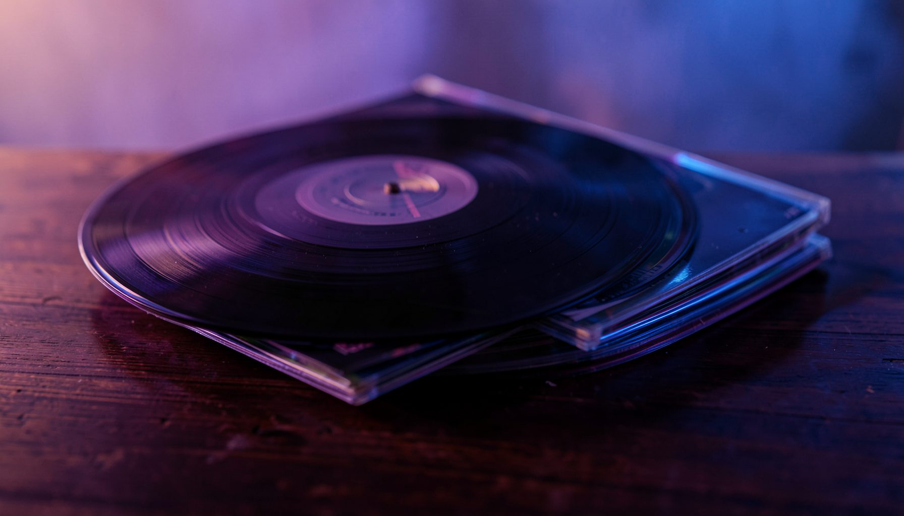 Pilha de discos de vinil e CDs sobre uma mesa de madeira escura, iluminados por luz quente e suave com reflexos em tons de roxo e azul